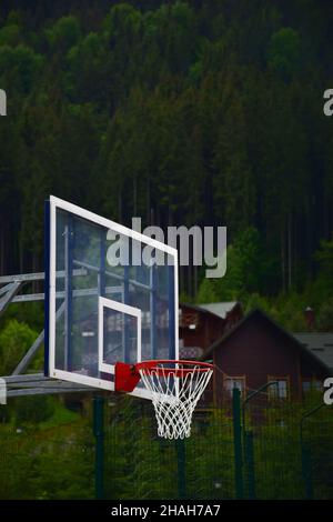 Basketball backboard and hoop with a net on a blurred background of wooden houses and green forest. Above is a place for an inscription Stock Photo