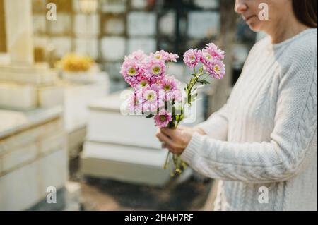 Crop woman with flowers in hands standing near gravestone in cemetery Stock Photo