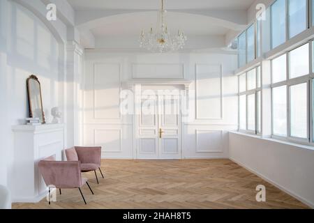 Interior of spacious white hall with armchairs placed near artificial fireplace with statuette and mirror on it opposite wide windows and big chandeli Stock Photo