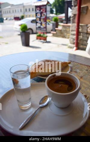 A cup of coffee, a croissant and a glass of water on a saucer are on a table in a roadside cafe. The background is very blurred Stock Photo