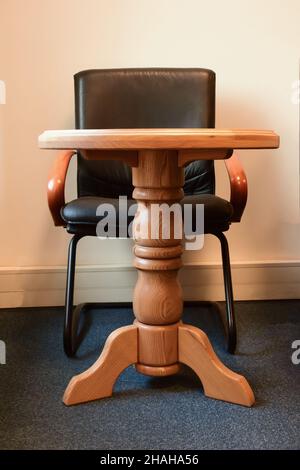 Decorative wooden table on one leg and behind it a black leather armchair against the background of the office wall Stock Photo
