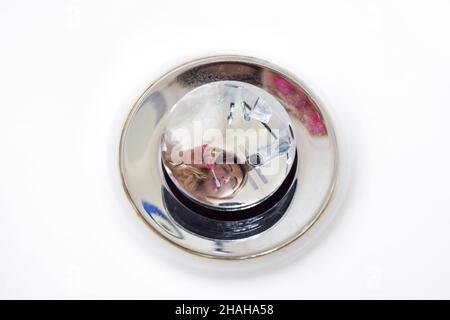 Reflection of a Caucasian woman brushing her teeth in a shiny stopper to stop the drain of water in the sink. Toothbrushes and toothpaste are also ref Stock Photo