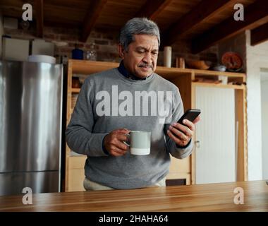 Multi-cultural elderly male reading daily news on smartphone while standing in modern kitchen. Stock Photo