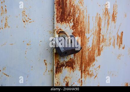 A black padlock hangs on an iron closed gate with peeling paint and rust Stock Photo