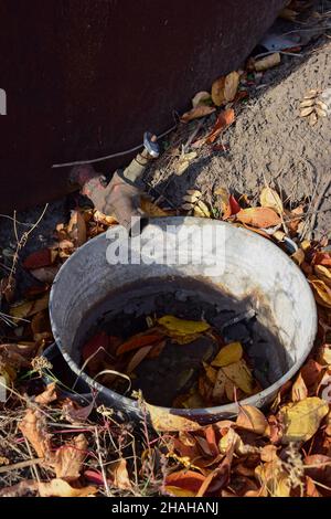 A closed water tap on the street, and under it is a rusty and leaky container without water and dry leaves in it. Stock Photo