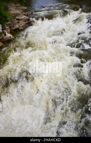 A stormy stream of a mountain river with a foamy waterfall flows between the rocky banks Stock Photo