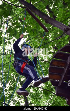 Young teen boy climbs hanging stairs from one tree to another in a rope amusement park Stock Photo