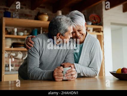 Happily retired elderly multi-cultural couple smiling, heads together. Wife holding husband in modern kitchen.  Stock Photo