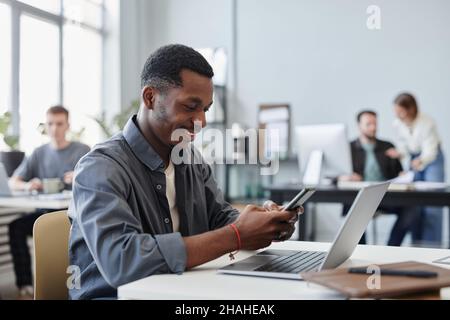African young businessman typing a message on mobile phone and smiling while sitting at his workplace with laptop at office Stock Photo