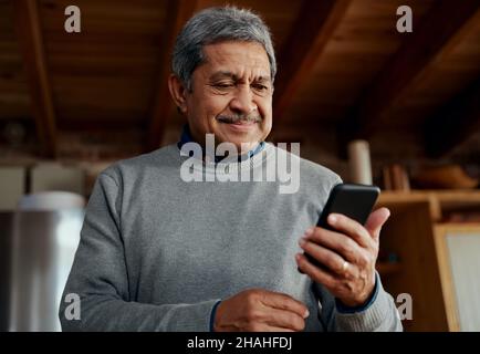 Smiling multi-cultural elderly male reading a message on smartphone while standing in modern kitchen. Stock Photo