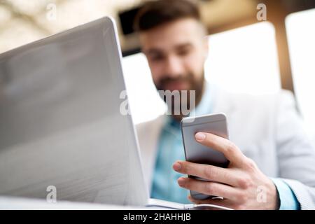 Handsome bearded man in stylish white and blue suit works on computer checking emails on smart phone. Stock Photo