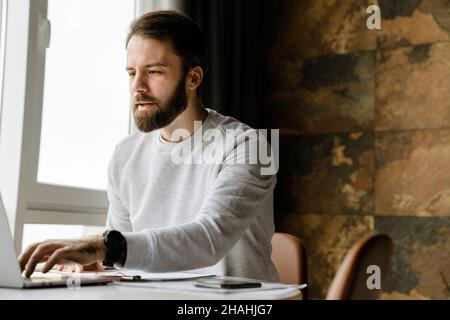 Bearded man working with laptop and papers while sitting at table indoors Stock Photo