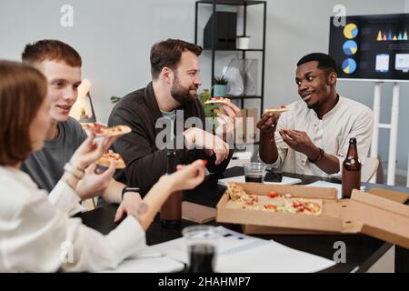 Business colleagues eating pizza at the table and talking to each other during lunch at office Stock Photo