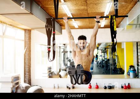 Fitness man hanging on horizontal bar performing legs raises, in the gym. Stock Photo