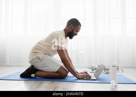 Smiling young black bearded man sit on mat on floor in room with dumbbells, bottle of water, watch workout Stock Photo