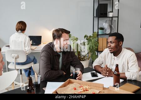 Two businessmen sitting at the table with pizza and alcohol drinks and talking to each other after work at office Stock Photo