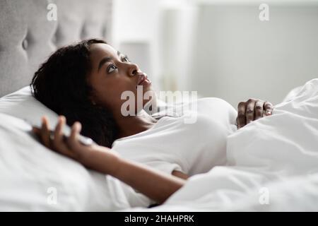 Unhappy black woman laying in bed, staring at the ceiling Stock Photo