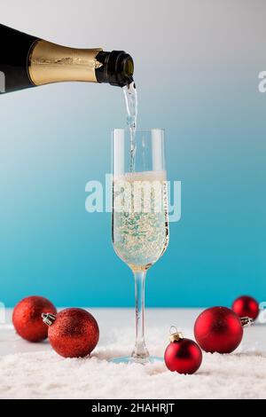 A tall glass is pouring champagne from a bottle on artificial snow with Christmas balls on a blue background. Stock Photo