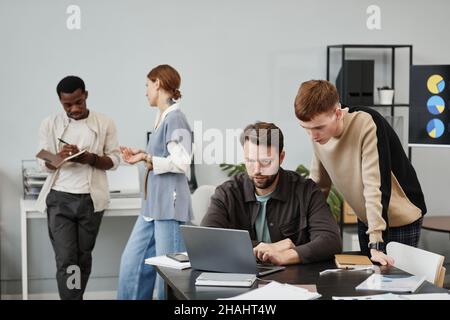 Two designers using laptop to create new project together at the table with other colleagues in the background at office Stock Photo