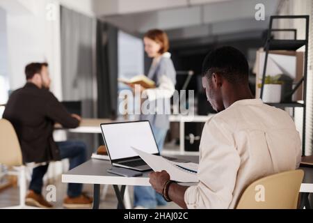 Rear view of African businessman sitting at his workplace in front of laptop and reading documents, he working at office with his colleagues Stock Photo