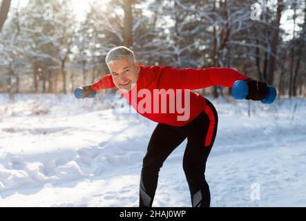 Fit senior sportsman with dumbbells doing powerlifting exercises in snowy park. Outdoor winter strength training Stock Photo