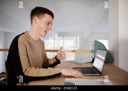 Young man smiling and talking online with his colleague on laptop while sitting at the table at office Stock Photo