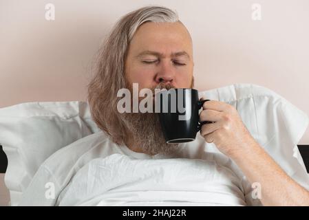 Bearded man enjoying his morning coffee in bed to cheer up before starting new day. Closeup Stock Photo