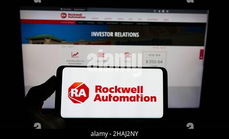 Person holding mobile phone with logo of American company Rockwell Automation Inc. on screen in front of business web page. Focus on phone display. Stock Photo