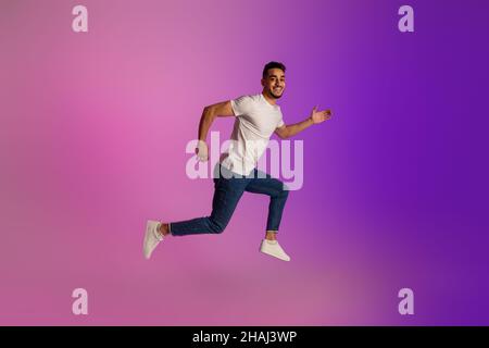 Full length portrait of positive young Arab man in casual wear jumping in neon light, copy space Stock Photo