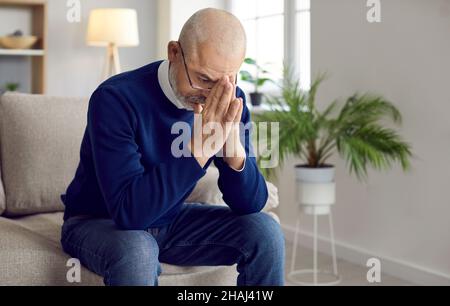 Stressed, worried senior man sitting on couch, praying to God and asking for help Stock Photo