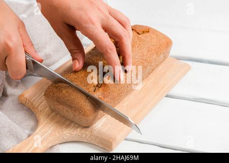 Woman hands holding knife and cutting sourdough homemade bread on cutting board on white wooden background Stock Photo