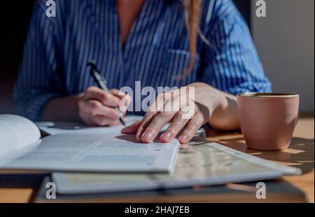 Student hands closeup writing with pen in notepad, taking notes from book, studying with textbook in cafe with coffee cup. Stock Photo