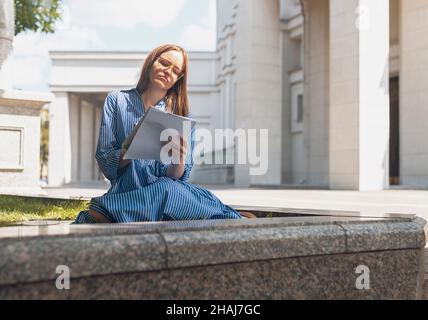Female student sitting on grass with notebook, writing smth, taking notes near university building. Modern woman learning with book. Stock Photo