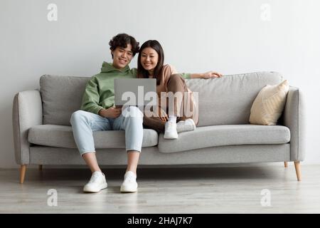 Happy young Asian couple using laptop, relaxing together on sofa at home, copy space Stock Photo