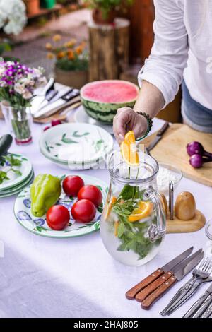 Woman hand putting slice of orange and mint herb into cold lemonade in carafe. Preparing cold drink for garden party Stock Photo