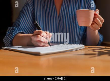 Student hands writing with pen in notepad, taking notes and holding coffee cup in hands. Stock Photo