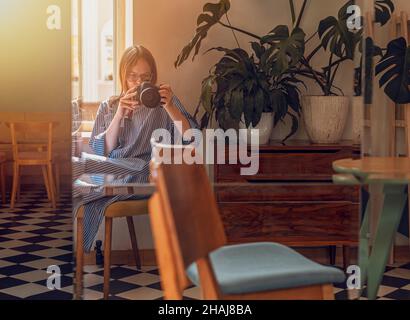 Young woman taking photo of herself, her reflection in mirror in modern cafe with day light and plants. Photographer with professional camera. Stock Photo