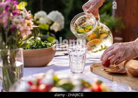 Pouring water with mint herbs and lemon from pitcher into drinking glass. Woman preparing healthy food and drinks Stock Photo