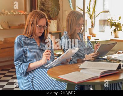 Young woman in eyeglasses in modern cafe with books and notebooks, learning and preparing for exam. Stock Photo
