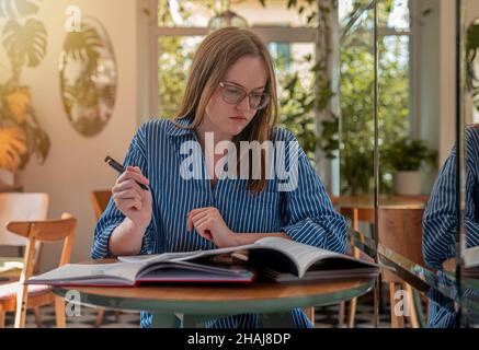 Student in eyeglasses in modern cafe with books and syllabus, studying in trendy coffee shop with day light. Stock Photo