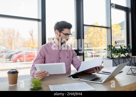 Businessman looking at papers and holding digital tablet near devices and coffee to go in office Stock Photo