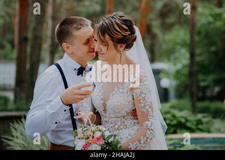 bride and groom with glasses of champagne in the park celebrate their wedding day. A loving couple of newlyweds drinks champagne, hugs and kisses Stock Photo