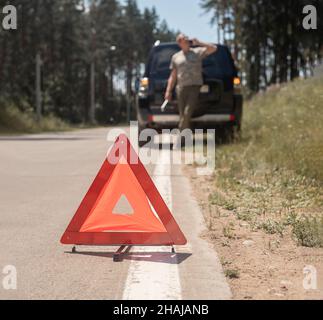 Red triangle caution sign on road after car break down and blurred driver speaking on cellphone, calling emergency repair service. Stock Photo
