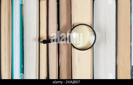 Magnifying glass over stack of books with many pages. Concept of search and investigation in library. Stock Photo