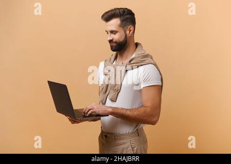 Smiling positive man holding laptop in hands and typing. Blogger making posts in social networks, chatting with followers. Indoor studio shot isolated on orange background Stock Photo
