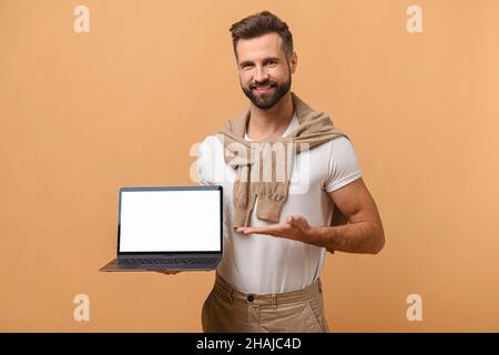 Positive smiling man freelancer with beard showing and holding laptop with empty screen, satisfied with teleworking, likes his job. Indoor studio shot isolated on orange background, mock-up Stock Photo