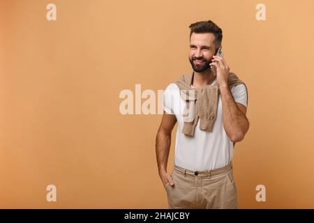 Young adult man with beard talking phone, calling to discuss plans, looking away with dreamy facial expression. Indoor studio shot isolated on orange background Stock Photo