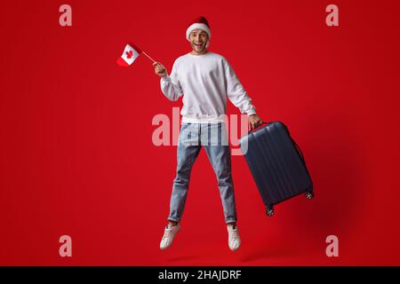 Man Wearing Santa Hat Jumping In Air With Canadian Flag And Suitcase Stock Photo