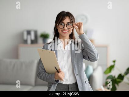Successful psychologist. Portrait of arab female psychologist holding clipboard, smiling at camera in modern office Stock Photo