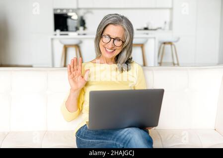 Video call concept. Happy senior woman using laptop computer for video connection from home, mature female looks at webcam and greeting colleagues, waving and smiles sitting on sofa at home Stock Photo